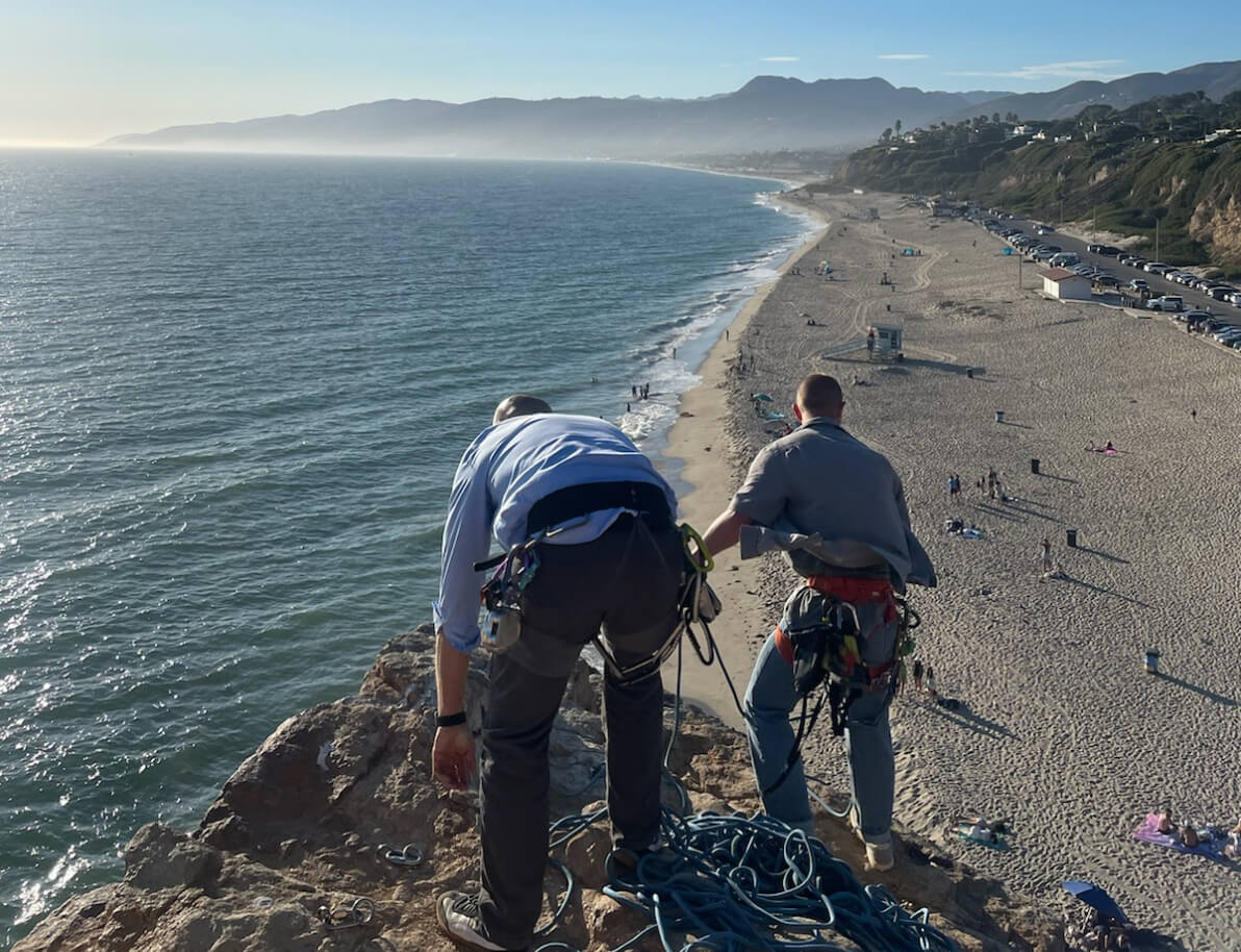 Two men standing at the top of a shoreside cliff with climbing gear